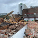 A demolished building with crumbling red brick and wooden beams in the foreground. A standing building with white sides and grey roof remains in the background.